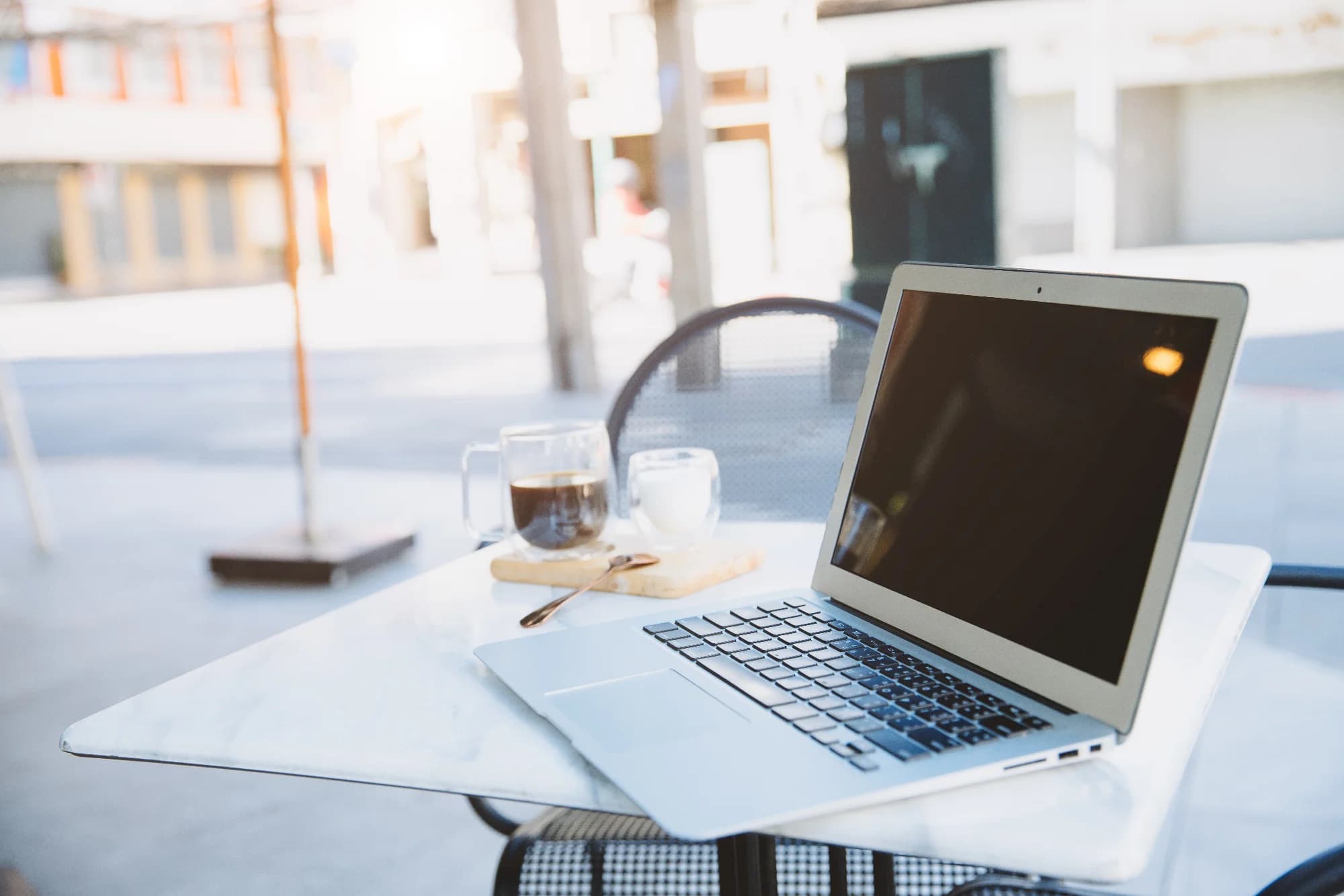 Un portátil abierto sobre una mesa de mármol claro en la terraza de un café. Una taza de cristal con café oscuro y un pequeño vaso de leche descansan sobre una tabla de madera junto a una cuchara. La luz del sol entra por la izquierda, iluminando la escena.